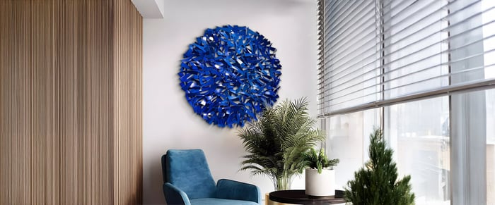 Contemporary blue metal 3D wall art with intricate overlapping elements, featured in a cozy corner with a blue chair, wooden paneling, and indoor greenery.