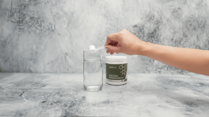 A hand enters from the right side of the frame scooping creatine into a glass with water.  The product is positioned to the right of the glass and the environment looks to be marble, the environment invites the notion that creatine is for everyone.