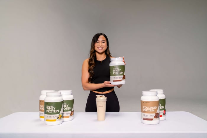 A young healthy woman standing behind a table with Grass-Fed Whey Protein arranged on it