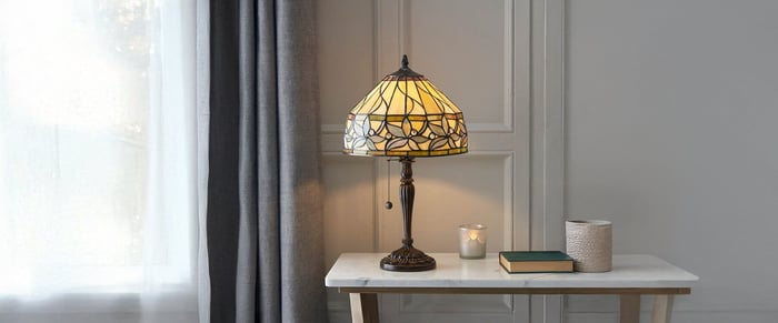 Tiffany-style table lamp with a yellow stained glass shade sits on a white console by a window with sheer curtains and grey drapes.