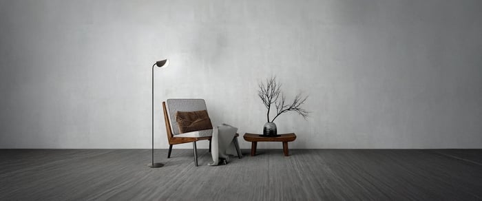 A modern floor lamp next to a cushioned chair with a brown pillow and a gray blanket, paired with a low wooden table and a decorative branch in a vase, against a textured wall.