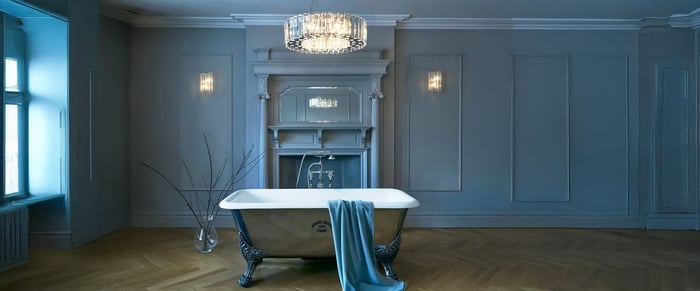 A modern bathroom lights featuring a freestanding white bathtub against a dark accent wall. Multiple wall-mounted light fixtures and ceiling lights provide soft ambient lighting, complementing the grey tiled wall's elegant texture.