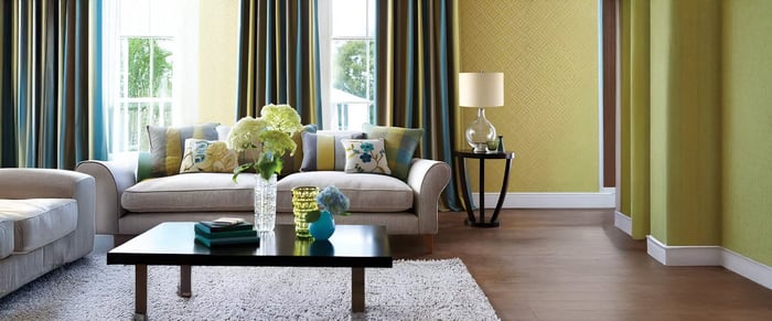 A cozy living room featuring a light grey sofa adorned with colorful throw pillows, a black coffee table with decorative vases, and a modern table lamps on a round side table, complemented by striped curtains and patterned wallpaper.