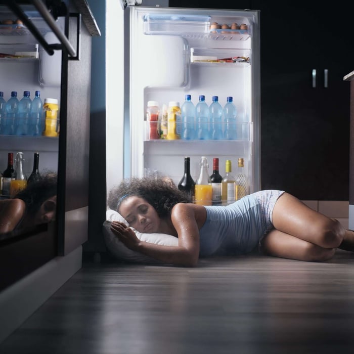 Woman lying on a kitchen floor in front of an open refrigerator, seeking relief from menopause night sweats and nighttime overheating.