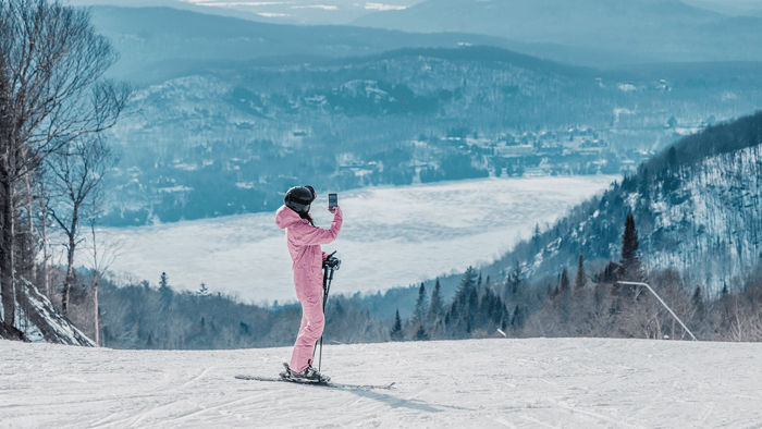 Woman in a pink ski suit standing on a snowy slope, holding ski poles and a smartphone, with a mountain and frozen lake view, wears engagement ring.
