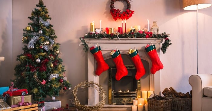 Christmas decoration ideas featuring a fireplace with candles and pine garland on the mantle, red socks hanging, candles on the floor next to baskets of chopped wood and pinecones, a decorated Christmas tree, and a wagon filled with gifts and toys