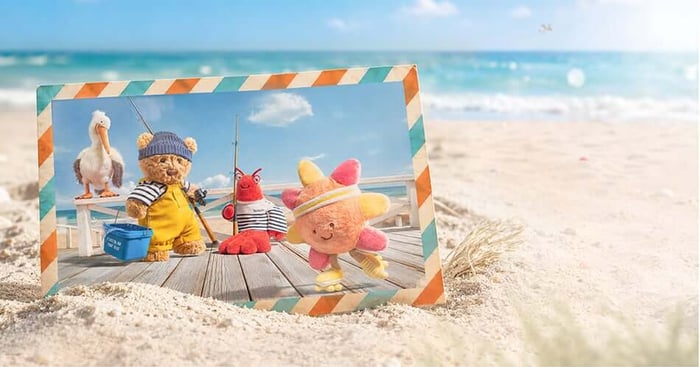 Bartholomew Bear Seafarer Outfit and friends pose on a seaside pier inside a postcard frame with sand and ocean behind.