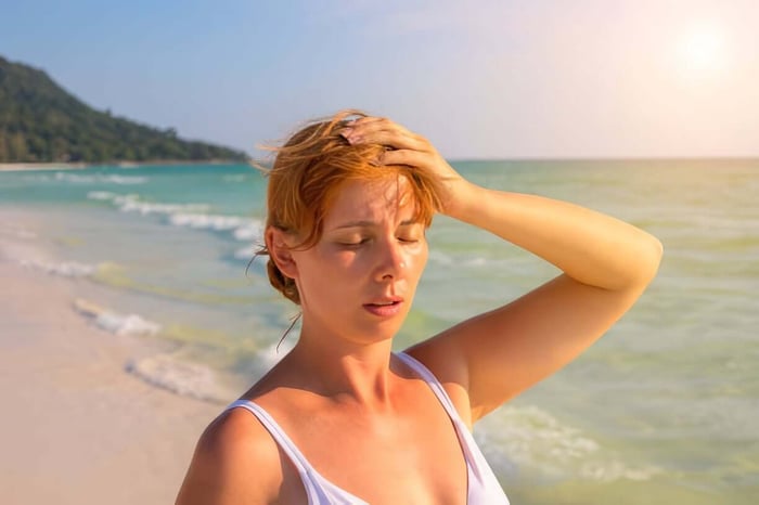 Woman standing on a sunny beach holding her head, showing discomfort from heat-induced migraine