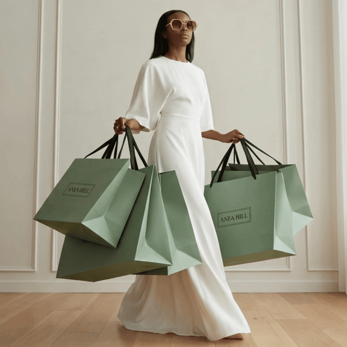 Woman in a flowing white dress carrying multiple green ANEA HILL shopping bags, wearing pink designer sunglasses, styled as the best sunglasses for UV protection in a clean, minimal luxury setting