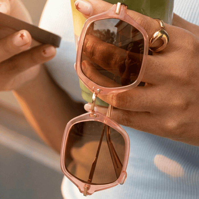 Close up of a woman holding pink Anea Hill sunglasses and an iced green drink while using her phone in warm golden hour light, showcasing the best sunglasses for women in a bright, airy lifestyle moment