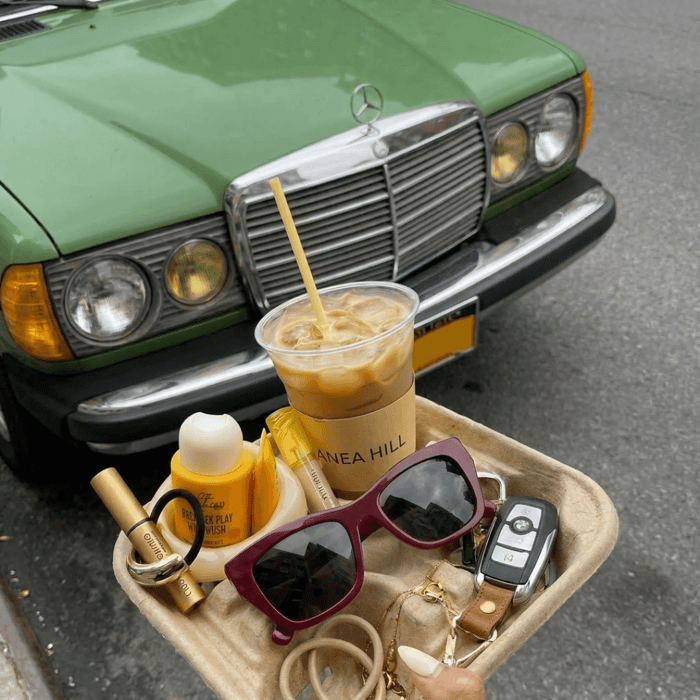 Best travel sunglasses for women: ANEA HILL Chablis frames in deep maroon styled in a travel tray with car keys, jewelry, and an iced coffee in front of a vintage green Mercedes.