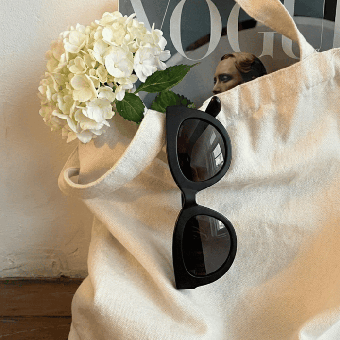 Cat eye sunglasses for women styled on a canvas tote with white hydrangeas and a magazine, featuring bold black frames in a soft, neutral setting.
