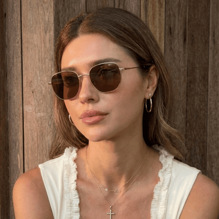 Woman wearing gold metal aviator sunglasses for small faces, with brown lenses, seated against a wooden backdrop in warm natural light, styled in a cream ruffled top with layered gold necklaces and hoop earrings