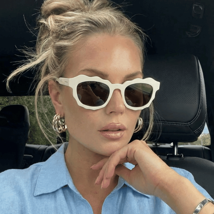Woman wearing white acetate sunglasses with brown polarized lenses while sitting in a car, styled with gold hoop earrings and a blue linen shirt, illustrating polarized lenses vs non for everyday driving wear.