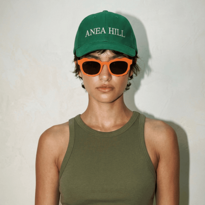 Model wearing ANEA HILL green cap and bold orange scallop sunglasses for women with a modern scalloped frame and dark lenses, styled with an olive tank top against a clean neutral background.
