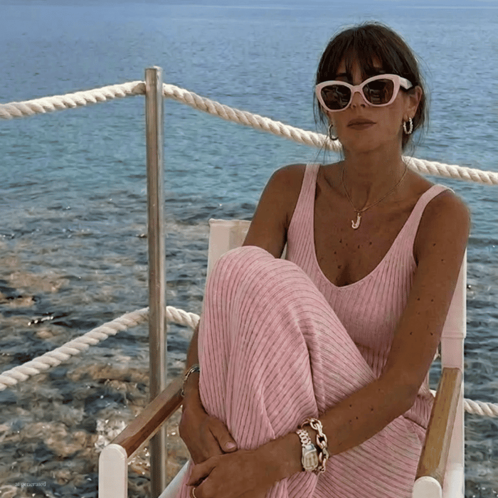 Woman seated by the sea wearing blush pink small frame sunglasses, paired with a soft pink knit dress and gold jewelry. Coastal Mediterranean setting with clear blue water, white rope railing, and natural sunlight, showcasing elegant small frame sung