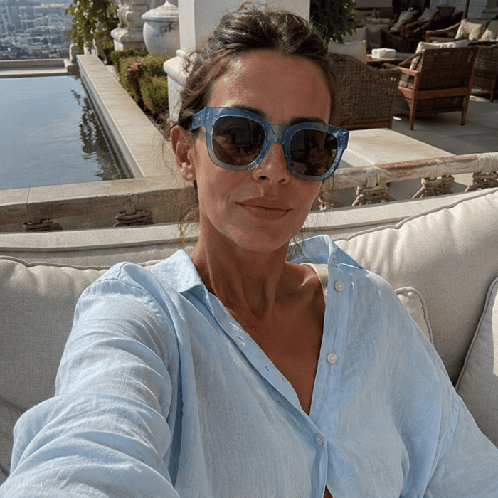 Woman relaxing at a luxury resort terrace, taking a selfie while wearing translucent blue Summer Travel Sunglasses. She is seated on a cream outdoor sofa overlooking a reflecting pool and elegant hotel architecture, dressed in a light blue button dow