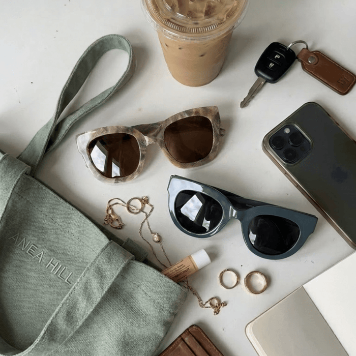What luxury sunglasses are worth buying for everyday use: two ANEA HILL frames styled alongside an iced coffee, car keys, phone, and ANEA HILL tote bag showing how they belong in your daily life.