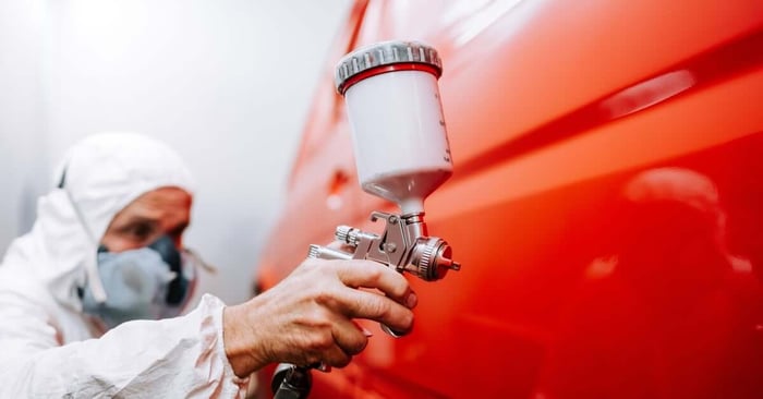 A man in a white protective suit and mask using a spray gun with a white tank to paint a red vehicle.