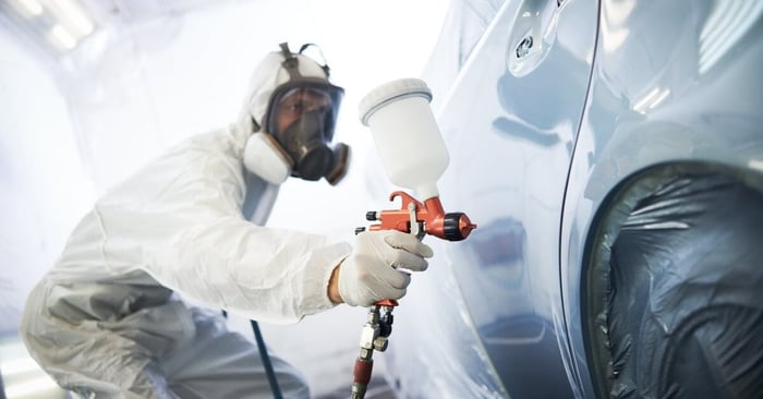 A person in a white protective suit and black respirator uses a spray gun with a hose to spray paint a vehicle.