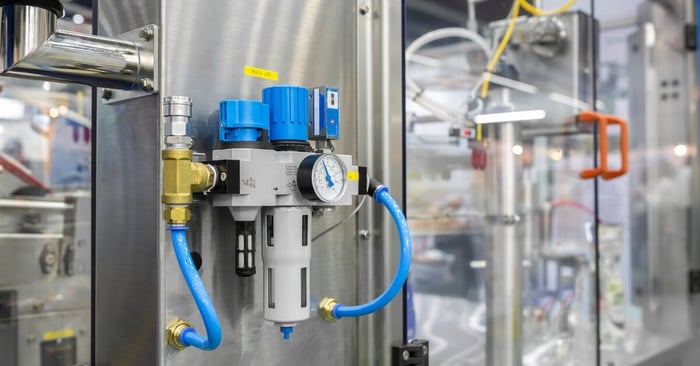An industrial pneumatic system with blue hoses, stainless steel panel, pressure gauge, and blurred machinery behind glass.