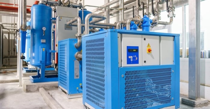 Blue industrial air dryers and tanks with control panels and gauges connected by metal piping in a clean facility.