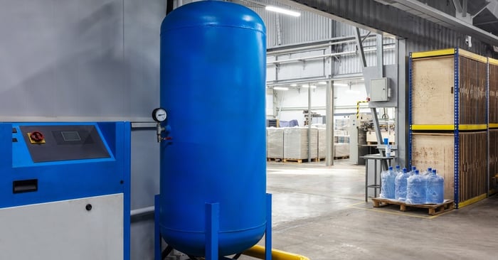 A blue vertical air receiver tank connected to a compressor in a warehouse, with pallets and storage racks nearby.