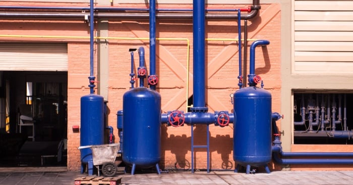 Blue industrial pressure tanks and piping system mounted outside a factory building with valves and connected pipework.