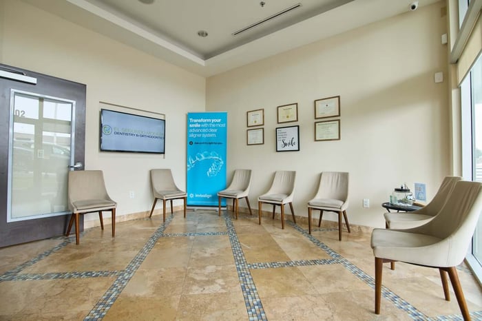 Dental office waiting room with spaced chairs, framed certificates on walls, tiled floor, and large windows.