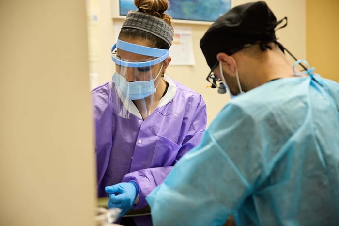 Dentist and dental assistant wearing protective gear while performing a dental procedure in a treatment room.