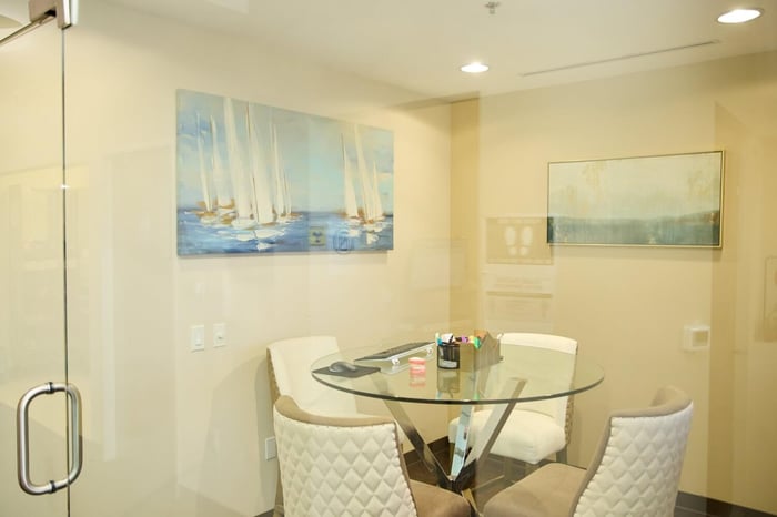 Private consultation room with glass table, white chairs, and coastal artwork inside a modern dental office.