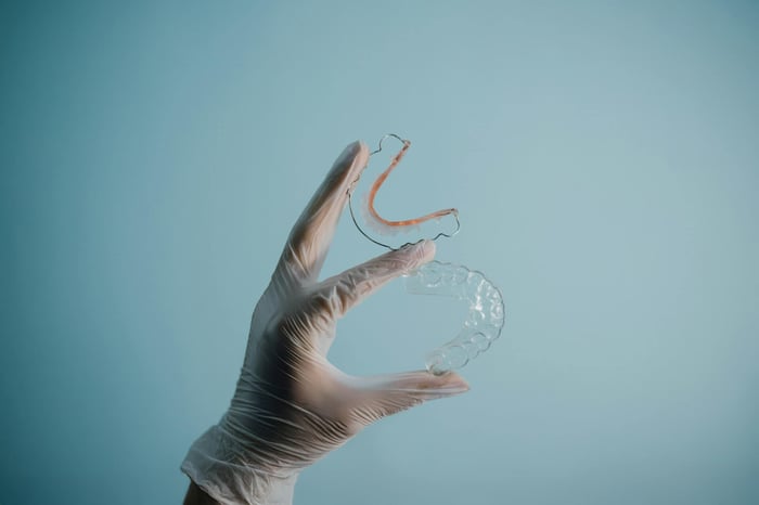 Gloved hand holding a clear dental retainer and orthodontic wire appliance against a light blue background.