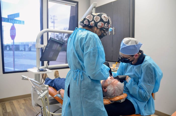 Two dental professionals wearing protective gowns and masks performing a dental procedure on a patient in an exam room.
