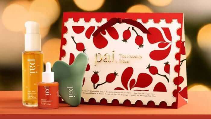 Pai Skincare Rosehip BioRegenerate Oil, serum bottle, and jade gua sha tool displayed with festive red botanical gift box against warm bokeh lights