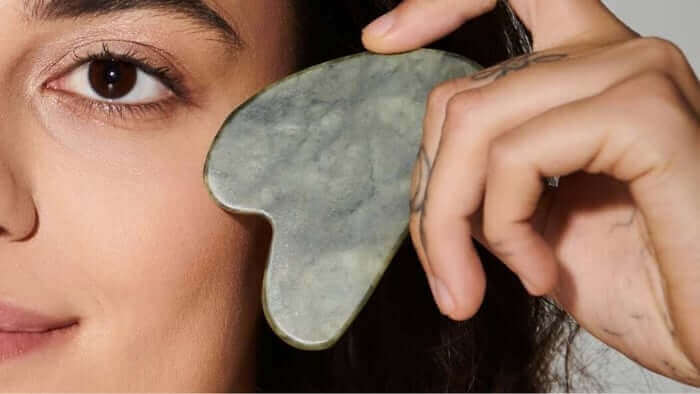 Woman using green jade gua sha stone on her cheek for facial massage and lymphatic drainage