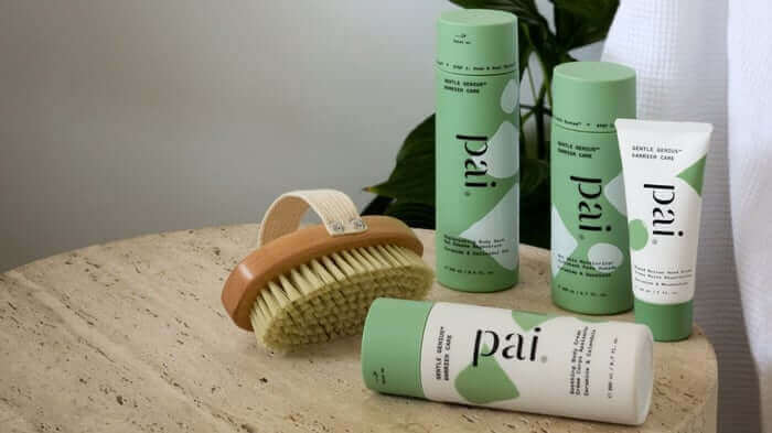 Pai Skincare body care collection with green and white packaging displayed on natural stone surface alongside wooden body brush
