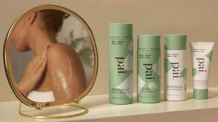 Pai Skincare ceramide body care collection with four green and white tubes arranged beside a round gold mirror reflecting a woman's shoulder