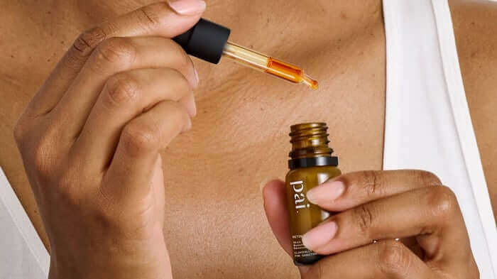 Hands applying golden facial oil from amber glass bottle with dropper onto chest, gentle skincare routine for sensitive skin