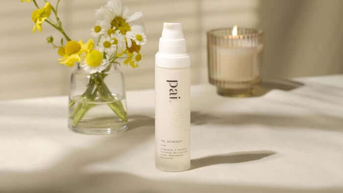Pai Anthemis Anti-Redness Serum bottle on marble surface with yellow wildflowers and lit candle creating a calming skincare routine scene