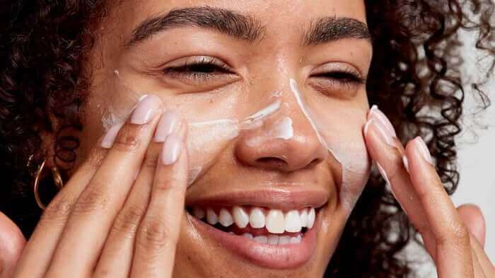 Smiling woman with curly hair gently massaging white cream cleanser onto her face with both hands