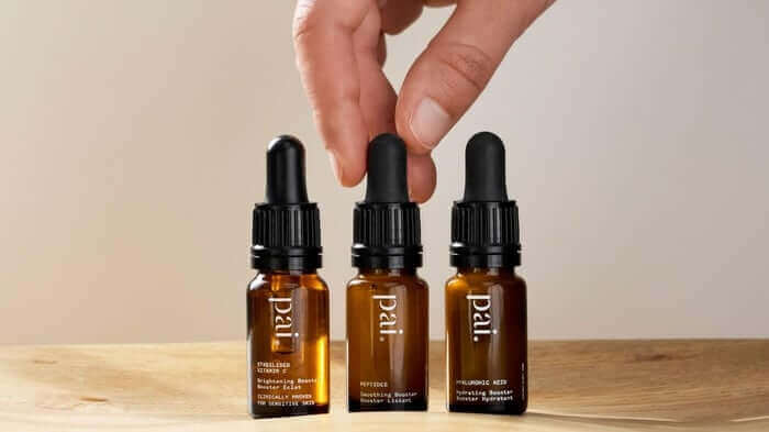 Hand reaching for three amber glass Pai skincare booster serums with dropper caps on wooden surface