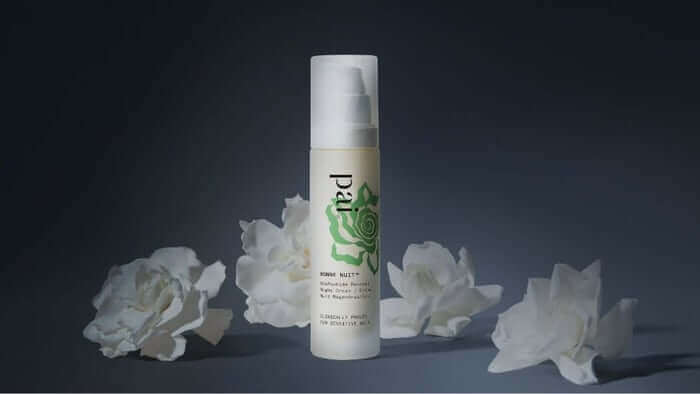 Pai Bonne Nuit Overnight Rebalancing Treatment bottle with white pump dispenser surrounded by delicate white rose petals on dark background