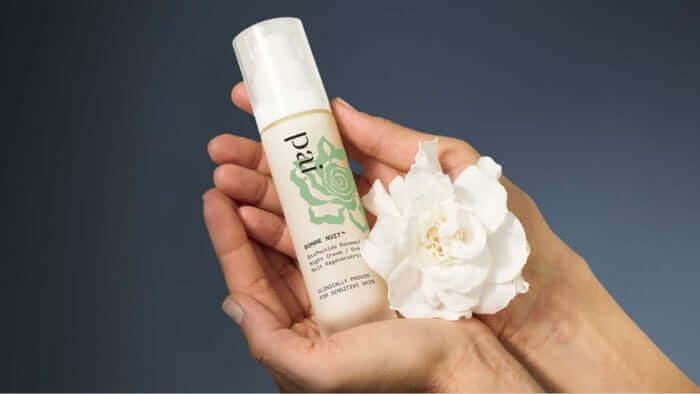 Hands gently holding Pai Bonne Nuit night cream with white flower against soft blue background