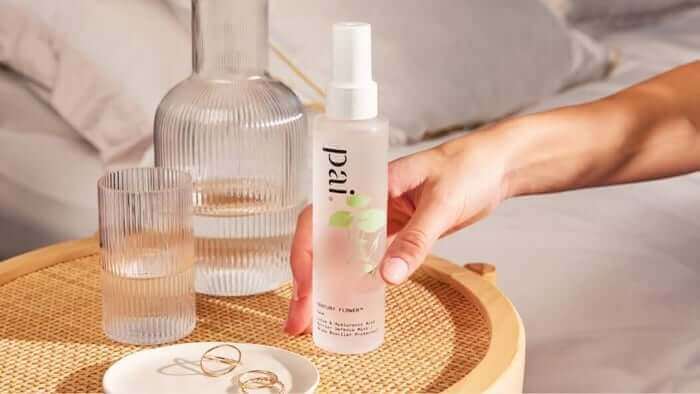 Hand holding Pai Hyaluronic Acid serum bottle with ribbed glass vase and jewelry on woven tray