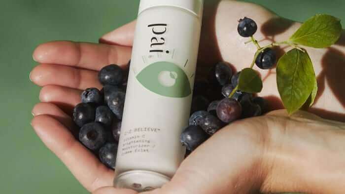 Hands holding Pai Skincare product bottle surrounded by fresh blueberries and green leaves against soft green background