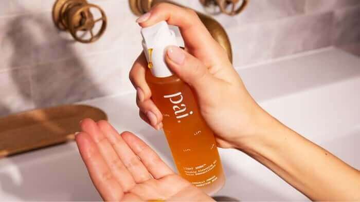Hand dispensing Pai rosehip oil from amber glass bottle into palm in bathroom setting for glowing skin routine