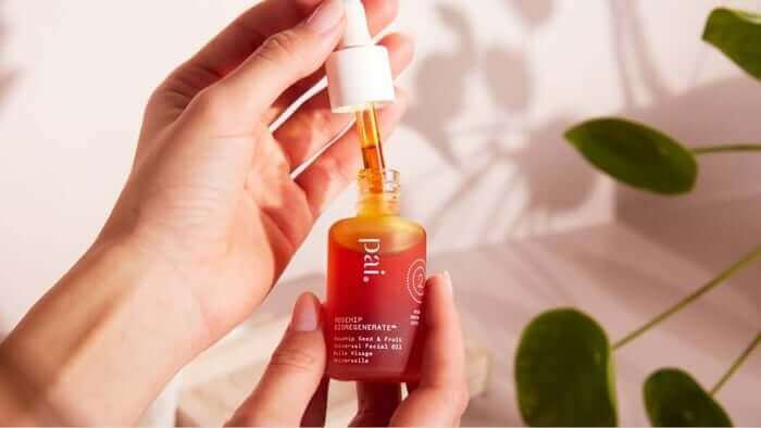 Hand holding Pai Rosehip BioRegenerate Oil bottle with dropper, showing the golden-orange facial oil against soft natural lighting with green leaves