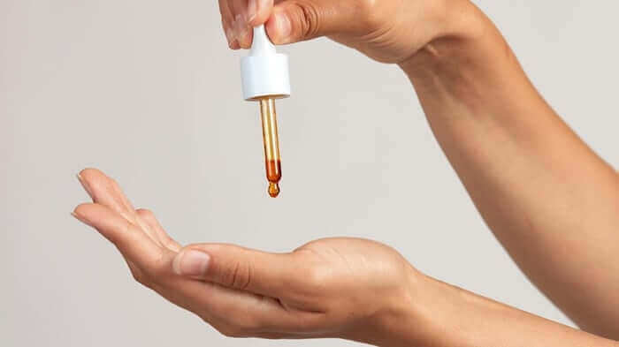 Hand dispensing golden face oil from white dropper bottle onto open palm