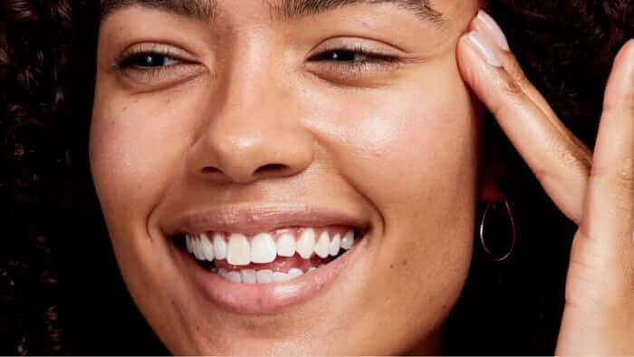 Smiling woman with curly hair touching her temple area, showing clear glowing skin