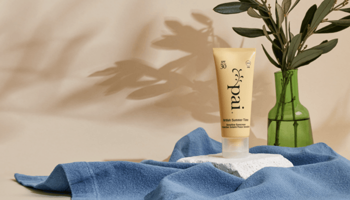 Pai British Summer Time SPF 30 sunscreen tube with golden packaging on blue linen fabric beside green glass vase with eucalyptus